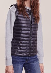 Padded vest in dark navy, featuring a smooth, shiny texture, zip closure, and side pockets, worn over a light gray long-sleeve shirt.
