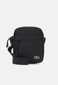 Černá crossbody taška vyrobená z odolné tkaniny, se zipovým uzavíráním, nastavitelným popruhem a malým zeleným krokodýlím logem na přední straně.