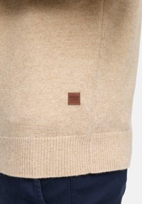 Beige wollen trui met een zachte textuur, voorzien van een geribbelde zoom en een klein bruin logo label op de onderkant.
