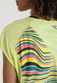 Primer plano del hombro de una mujer que lleva una blusa amarilla con un panel de rayas onduladas multicolores, con la cinta de un sujetador deportivo negro visible.