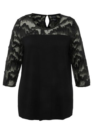 Haut noir avec un devant uni et des manches en dentelle noire transparente au motif ondulé. Détail de dos en forme de goutte et col rond.