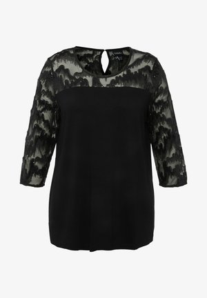 Top nero con davanti in tessuto solido e maniche in pizzo nero trasparente con un motivo ondulato. Dettaglio con apertura a chiave sulla schiena e scollatura rotonda.