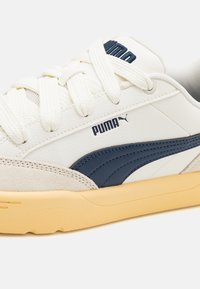 Bílé a tmavě modré tenisky s hladkým koženým svrškem, semišovými akcenty, silnou gumovou podešví a tkaničkami s tkaním, na kterých je logo Puma.