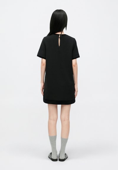 Mujer de pelo largo y negro de espaldas, vestida con un vestido corto negro, calcetines grises y zapatos negros tipo slip-on, sobre un fondo blanco.