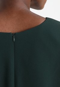 Dunkelgrüner Kleiderstoff mit glatter Textur, ausgestattet mit einem dezenten Reißverschluss auf der Rückseite. Schlichter, sauberer Ausschnitt und Design.