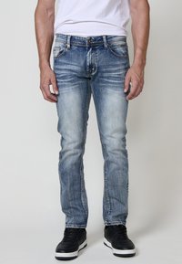 Jeans de mezclilla azul con un corte ajustado, áreas ligeramente desgastadas y diseño clásico de 5 bolsillos, combinados con zapatillas negras sobre un fondo neutro.