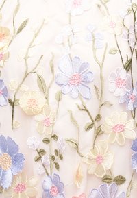 Broderie florală delicată în nuanțe pastel, cu flori mov deschis, galbene și roz și tulpini verzi pe un fundal transparent de pânză palidă.