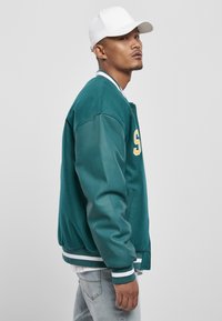 Teal bomberjack met leren mouwen, geribbelde manchetten en een geribbelde tailleband. Heeft een gele letter op de voorkant en een witte pet.