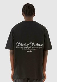 Tricou oversized negru, cu text alb pe spate, într-un font decorativ, detaliind fraze despre reziliență și stăpânire.