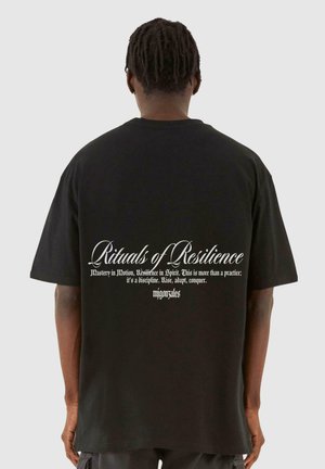 Sort oversized t-shirt med hvid tekst på ryggen i dekorativ skrifttype, der beskriver sætninger om modstandskraft og mestring.