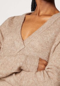 Beiger V-Ausschnitt-Pullover aus weichem, strukturiertem Stoff, mit gerippten Bündchen und einer lässigen Passform. Feine Nahtdetails entlang des Ausschnitts.