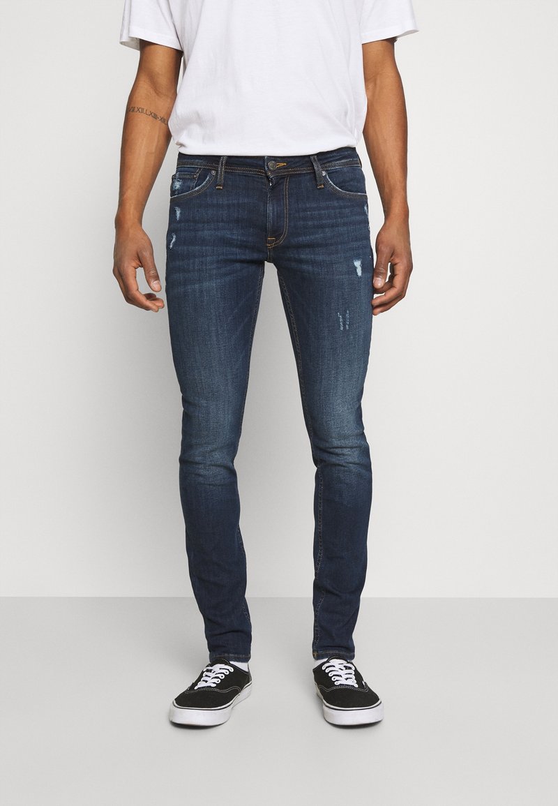 Homme portant un jean slim bleu foncé avec des détails délavés et usés, un t-shirt blanc et des baskets noires, debout devant un fond uni.
