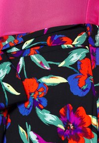 Jupe noire avec des motifs floraux rouge vif, violet et bleu portée avec un haut rose texturé, montrant les détails du tissu et de la fermeture éclair.