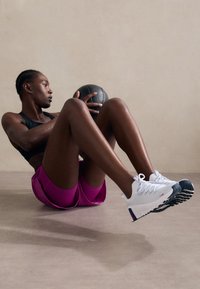 Svart idrettsbh, rosa treningsshorts med sideslisser og hvite sneakers med svarte såler. Individet holder en svart medisinball.