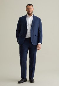 Marineblauw pak met een notched lapel en twee voorzakken, gecombineerd met een wit overhemd en zwarte schoenen. Gladde stof, getailleerde pasvorm.