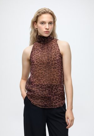 Femme blonde portant un haut marron sans manches à col montant imprimé léopard et un pantalon noir, debout avec une main dans la poche sur un fond uni.