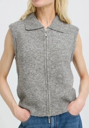 Mouwloze grijze gebreide vest met kraag en ritssluiting aan de voorkant, gedragen over een blauwe spijkerbroek door een persoon met de handen in de zakken.