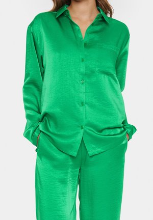 Kobieta ubrana w błyszczącą zieloną satynową koszulę zapinaną na guziki i pasujące luźne spodnie, dłonie w kieszeniach, jednolite jasne tło