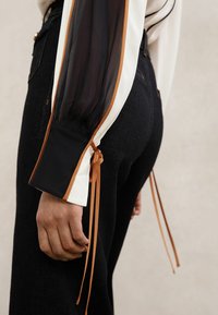 Pikkade varrukatega pluus, mis on valmistatud mustast läbipaistvast kangast valgete detailidega, mansettidel saab reguleerida pruuni värvi paeltega ning taljel on istuv must vöökoht.