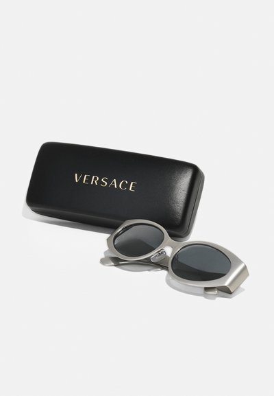 Versace saulės akiniai su kampuotais sidabriniais rėmeliais ir tamsiais lęšiais, padėti šalia juodos dėžutės su auksiniu "VERSACE" logotipu.