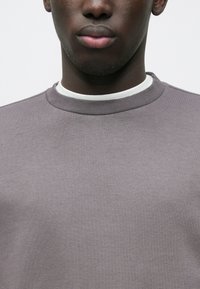 Gros plan sur la partie inférieure du visage et le cou d'une personne portant un pull gris à col rond par-dessus un T-shirt blanc, sur un fond uni.