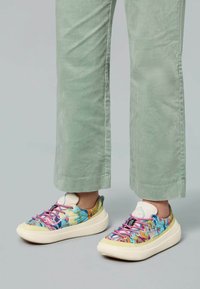 Kolorowe trampki z kwiatowym wzorem, posiadające białą gumową podeszwę, różowe sznurowadła oraz teksturowaną tkaninę. Noszone z jasnooliwkowymi spodniami sztruksowymi.