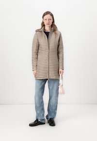 Beige Puffer-Jacke mit horizontaler Steppung, Reißverschluss vorne und Kapuze, kombiniert mit weiten blauen Jeans und schwarzen Schuhen, hält eine pinke Handtasche.