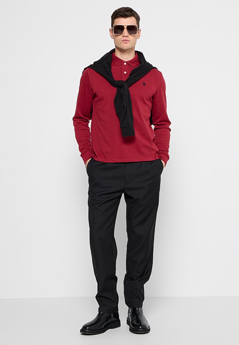 Polo-shirt rouge à manches longues, pull noir drapé sur les épaules, pantalon noir et chaussures noires. Le modèle porte des lunettes de soleil. Fond simple.