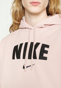 Sudadera rosa con un gran logo negro de "NIKE" con letras texturizadas y un pequeño swoosh negro. Hay cordones en el escote.