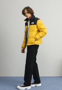 Gul og sort pufferjakke med lynlås foran og vandret quiltning, parret med sorte bukser og sorte sneakers med hvide såler.