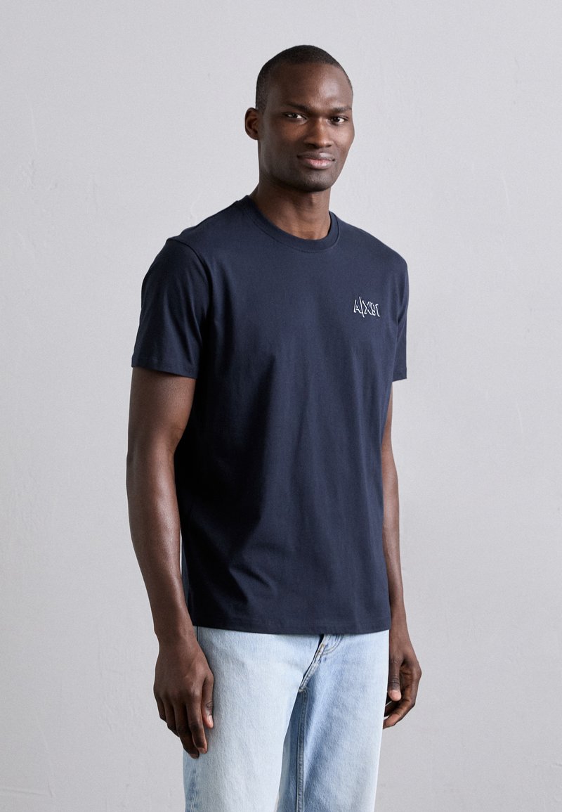 Camiseta de algodón azul marino de manga corta, con cuello redondo y un pequeño logo blanco en el pecho, combinada con jeans de mezclilla azul claro.