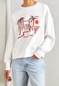 White sweatshirt featuring a red graphic print of palm trees and a sun, with ribbed cuffs and hem, worn with light blue jeans.