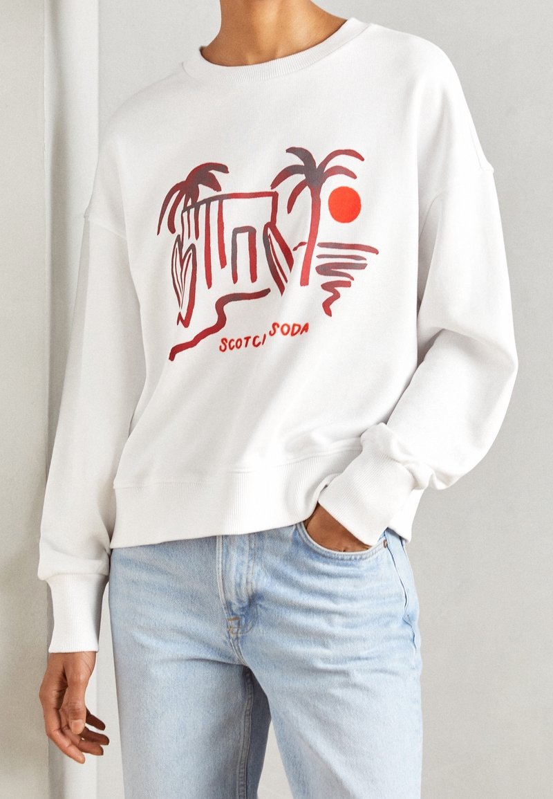 White sweatshirt featuring a red graphic print of palm trees and a sun, with ribbed cuffs and hem, worn with light blue jeans.