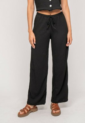 Femme portant un pantalon large texturé noir à taille coulissante, un haut court boutonné noir et des sandales marron à brides.