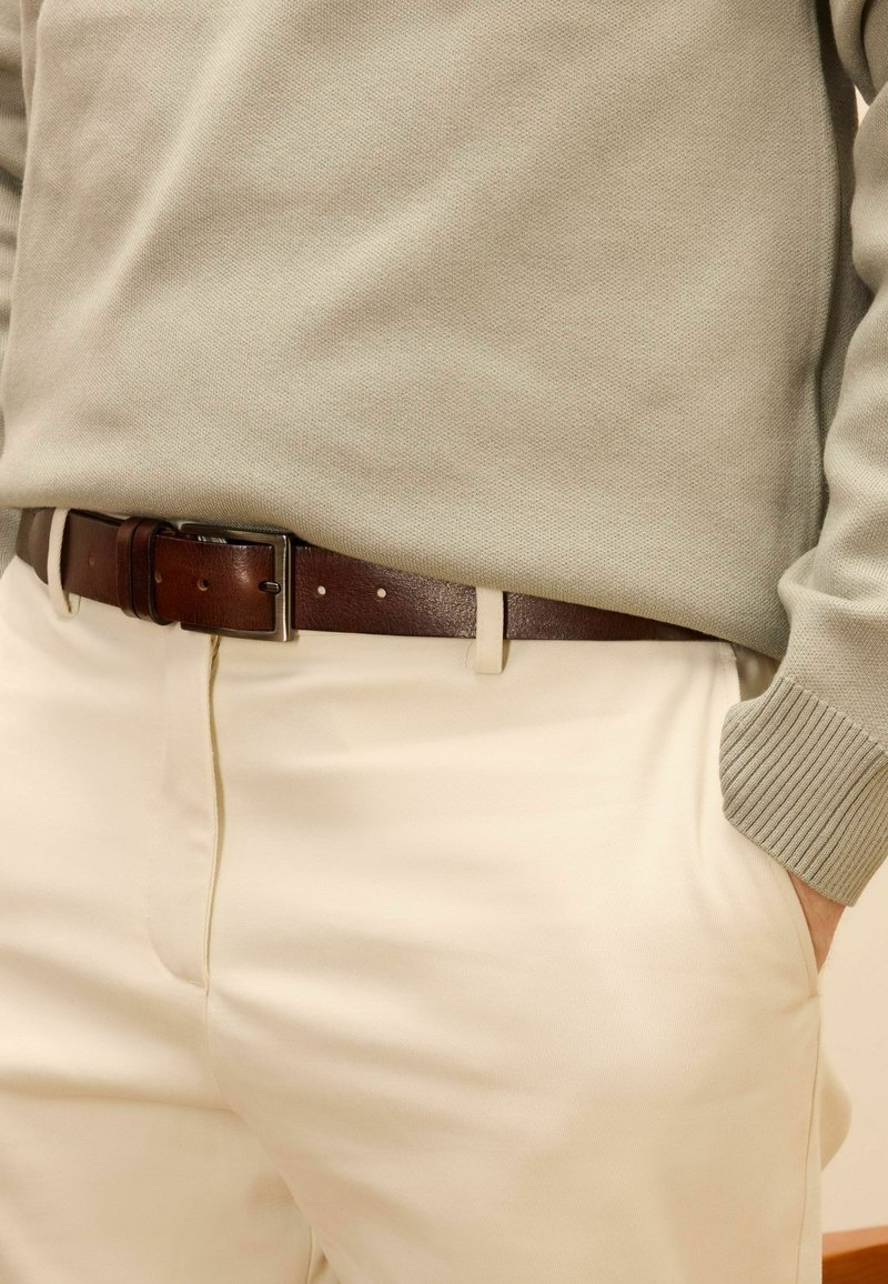 Lichtgrijze trui met geribbelde manchetten, bruine leren riem met zilveren gesp en crèmekleurige broek. Focus op textuur en kleurcontrast.