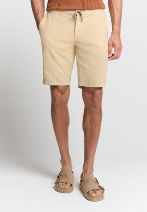 Shorts beiges en coton avec une taille élastique et un cordon de serrage. Dotés de poches latérales et d'une coupe décontractée. Portés avec des sandales marron.