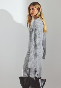 Gray ribbed knitted dress with long sleeves, featuring a loose fit. A black tote bag is held in one hand. Smooth texture and soft appearance.