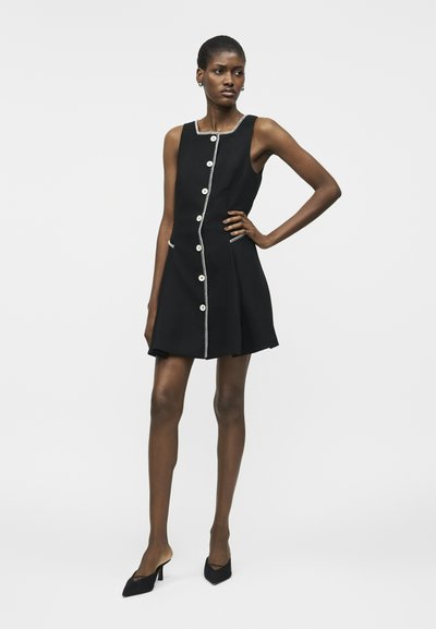 Γυναίκα με μαύρο φόρεμα χωρίς μανίκια, με λευκές λεπτομέρειες κουμπιών και περιμετρική ρέλι, στέκεται με το ένα χέρι στη μέση της φορώντας μαύρα παπούτσια με τακούνι.