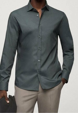 Mężczyzna w dopasowanej ciemnoszarej koszuli zapinanej na guziki oraz beżowych spodniach, trzymający w lewej ręce ciemny materiał lub ubranie.