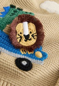 Beige neulepaita, jossa on teksturoitu leijonan kuvio, ruskea harja ja kirjailtuja yksityiskohtia keltaisessa, mustassa, sinisessä ja vihreässä. Sisältää leikillisiä kuvioita.