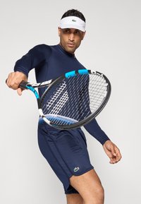 Camisa atlética de manga longa azul-marinho e calções com o logo Lacoste. Um jogador segura uma raquete de ténis azul e preta, usando uma viseira branca.