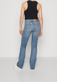 Persona che indossa jeans a vita alta dritti blu chiaro, top senza maniche nero e sneaker bianche, in piedi contro uno sfondo neutro.