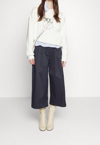 Vit oversized sweatshirt med grafisk design, lagd över en ljusblå randig skjorta, ihop med vida svarta byxor och krämfärgade ankelstövlar.