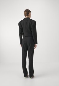 Black tailored suit with oversized shoulders, side zipper accents on pants, and a branded tag on the back. Smooth, structured fabric.
