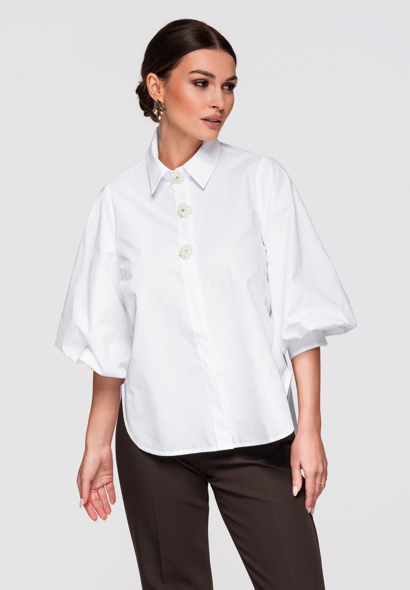 Femme portant un chemisier blanc à manches bouffantes avec des boutons floraux et un pantalon foncé, regardant vers sa gauche devant un fond uni.