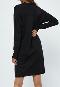 Una donna sta in piedi di spalle rispetto all'osservatore, indossando un vestito nero ampio con maniche lunghe. La sua mano è appoggiata sui fianchi.