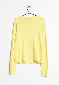 Žlutý top s dlouhými rukávy, volného střihu, vyrobený z lehké tkaniny. Má kulatý výstřih a boční rozparky na lemu pro lepší prodyšnost.