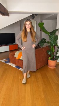 Žena s dlhými vlasmi v hnedom šaty stojí s rukami v bok v modernom obývacej izbe s veľkou rastlinou a oranžovou sedačkou.