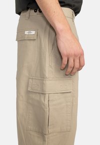 Beige cargo bukser med et tekstureret stof, der har en sidelomme og et mærke. Skåret med et lige ben-design.