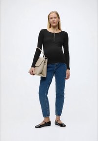 Γυναίκα με μαύρη μπλούζα μακρυμάνικη, μπλε τζιν παντελόνι και μαύρα φλατ παπούτσια, κρατώντας μια μπεζ τσάντα χειρός, στέκεται μπροστά σε απλό λευκό φόντο.